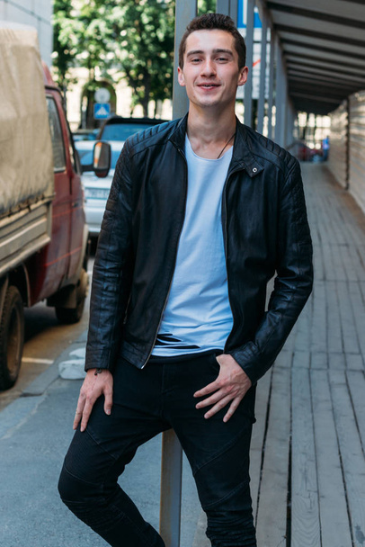attractive and handsome guy stands outside near the entrance and laughs in a white t-shirt and a black leather jackets. communication. - Foto, afbeelding
