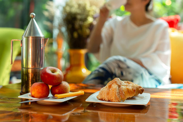 Bulanık kadın sabah kahvesini kruvasan ve ahşap masada elmayla sever.. - Fotoğraf, Görsel