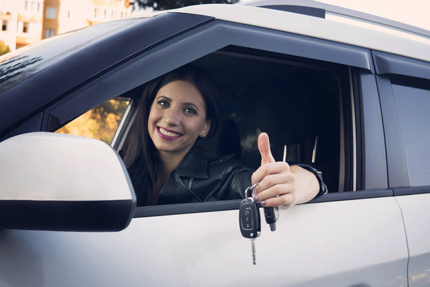 Beautiful car driver woman smiling showing new car keys and car. Caucasian girl sitting in automobile, smiling and demonstrating keys. Female owner of vehicle. Woman is happy to purchase auto. - Photo, Image