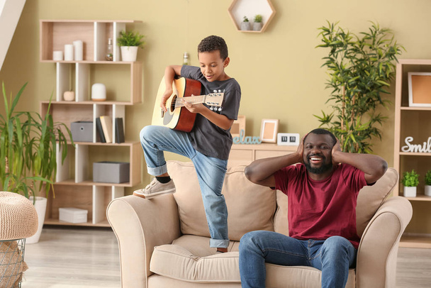 African-American man covering ears while his little son is playing guitar at home - Фото, изображение