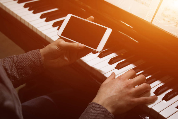 two male hands on the piano with a smartphone. palms lie on the keys and play the keyboard instrument in a music school. student learns to play. hands of a pianist. black dark background. - 写真・画像