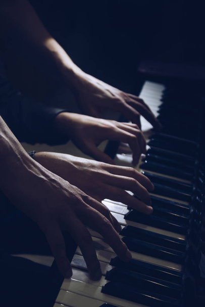 playing four male hands on the piano. palms lie on the keys and play the keyboard instrument in a music school. student learns to play. hands of a pianist. black dark background. - 写真・画像