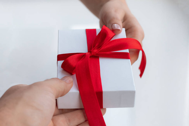 ギフトプレゼントボックスには、明るい白い背景に赤いリボンの弓が隔離されています。バレンタインデーのプレゼントを手から手に渡す。愛と休日の概念. - 写真・画像
