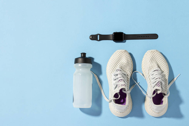 Fitness tracker, λευκά sneakers και ένα μπουκάλι νερό κάτω από το πρωινό φως του ήλιου σε μπλε φόντο.Jogging, τρέξιμο, fitness, cross fit, σκληρή προπόνηση. Πρωινό τρέξιμο. Μπάνερ. Επίπεδο lay, πάνω όψη - Φωτογραφία, εικόνα