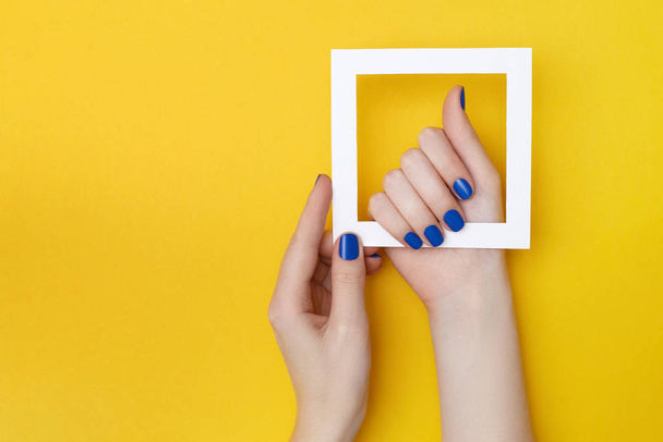 Vrouwelijke handen met manicure in trendy klassieke blauwe kleur met wit frame op de gele achtergrond - Foto, afbeelding