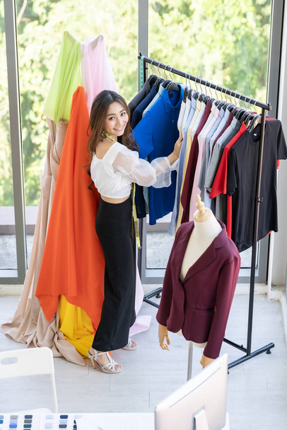 Top view of young adult fashion designer standing near clothing rack with colorful and fashionable stylish handmade clothes at her atelier studio as sole owner. Using for entrepreneur startup concept. - Foto, immagini