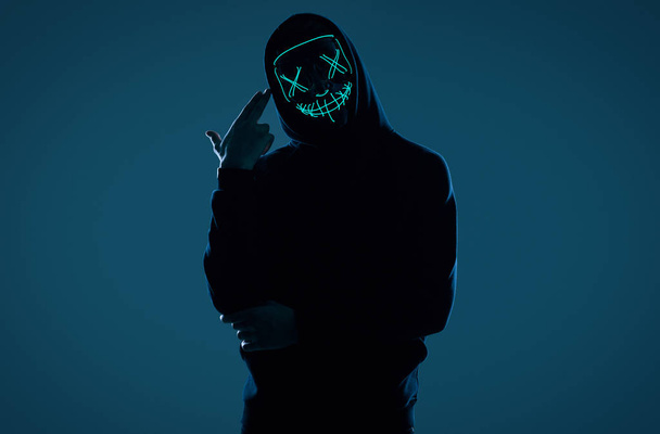 Anonymer Mann in schwarzem Kapuzenpulli versteckt sein Gesicht hinter einer Neonmaske - Foto, Bild