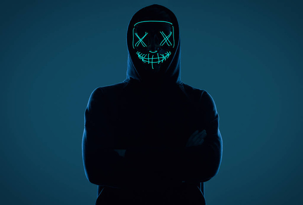 Siyah kapüşonlu isimsiz bir adam yüzünü neon bir maskenin arkasına gizliyor. - Fotoğraf, Görsel