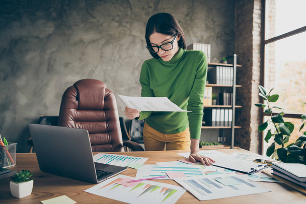 Photo of beautiful busy business lady notebook table paper stats on table analyzing money income standing near chair wear specs green turtleneck yellow pants modern office - Fotó, kép