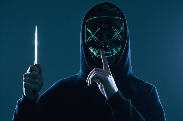 Hombre criminal anónimo con cuchillo de cocina en una sudadera con capucha negra y máscara de neón
 - Foto, Imagen