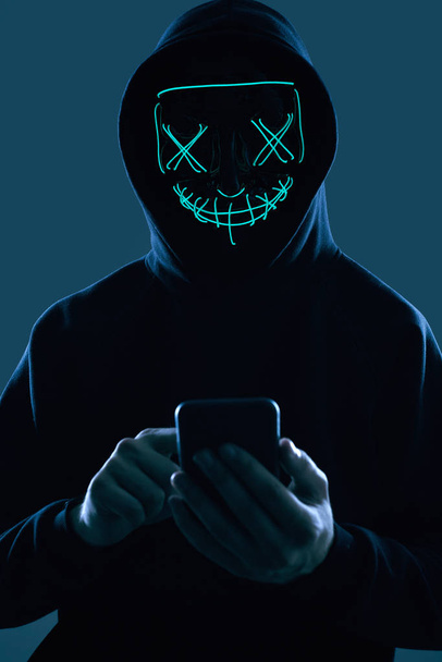 Anonymous man in a black hoodie and neon mask hacking into a smartphone - Photo, Image