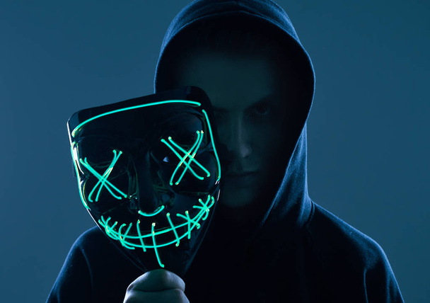 Anonimowy mężczyzna w czarnej bluzie chowa twarz za neonową maską. - Zdjęcie, obraz