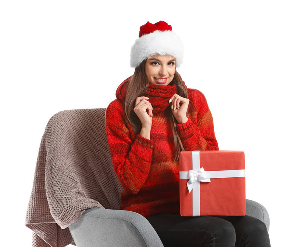 Beautiful young woman in Santa hat and with Christmas gift sitting in armchair on white background - Zdjęcie, obraz