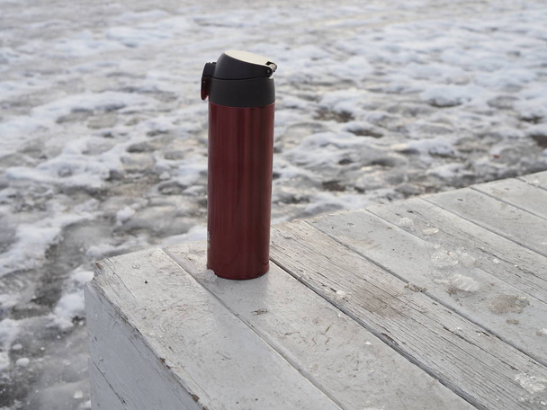 red thermocup on a white wooden table against the background of white snow in the chilly weather - 写真・画像