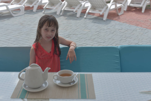 9-letnia dziewczynka pije czarną herbatę w parku wodnym lub na wakacjach przy basenie. Dziecko odpoczywa latem.. - Zdjęcie, obraz