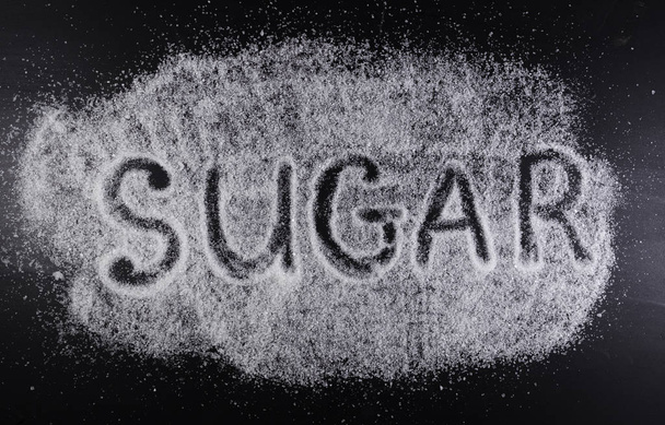 The word SUGAR on the spilled sugar on black background. Harm of sugar, diabetes disease medical concept. - Фото, изображение
