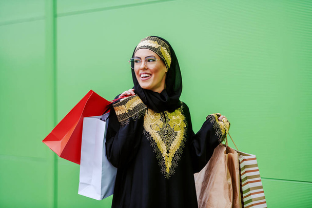 Giovane attraente donna araba ridente in usura tradizionale camminando all'aperto con borse della spesa in mano. Lei è soddisfatta del suo shopping
. - Foto, immagini