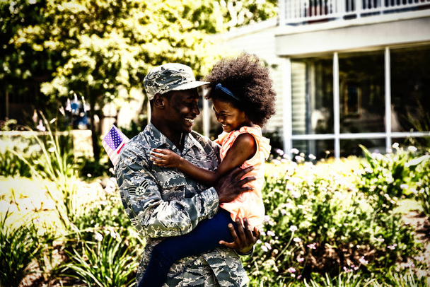 Side view of a young adult African American male soldier in the garden outside his home, holding his young daughter, both looking at each other smiling - Foto, Imagem