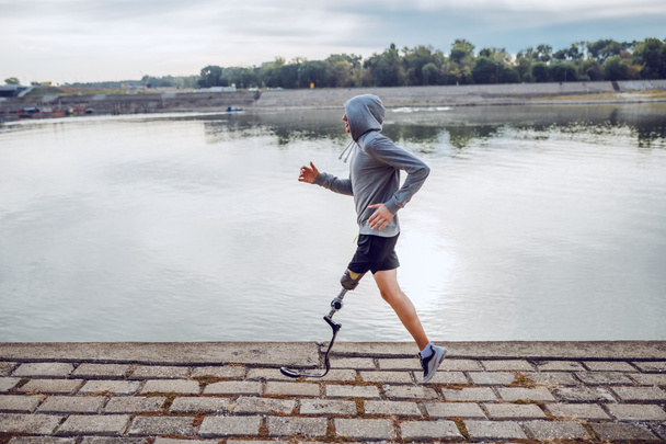 Side view of healthy caucasian handicapped sportsman in sportswear and with artificial leg running on quay. - Photo, Image