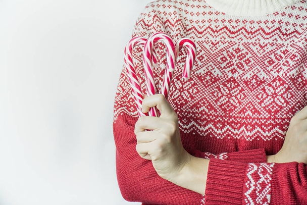 Κορίτσι σε ένα χριστουγεννιάτικο πουλόβερ κατέχει χριστουγεννιάτικα ζαχαρωτά μπαστούνια σε λευκό φόντο, close-up. Χριστουγεννιάτικο φόντο. Χώρος για κείμενο. - Φωτογραφία, εικόνα