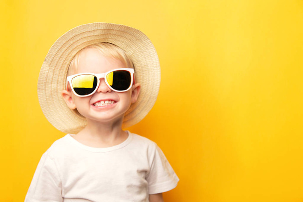 Portrait of a cute blond baby boy with a happy face smile in a straw hat and sunglasses on a yellow background looking at the camera. card with place for text - 写真・画像