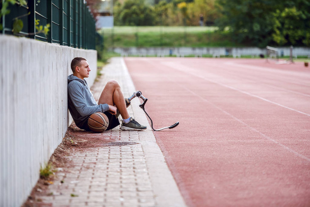 Side view of handsome fit serious sportive handicapped man in sportswear and with artificial leg sitting on racetrack with basketball ball. - 写真・画像