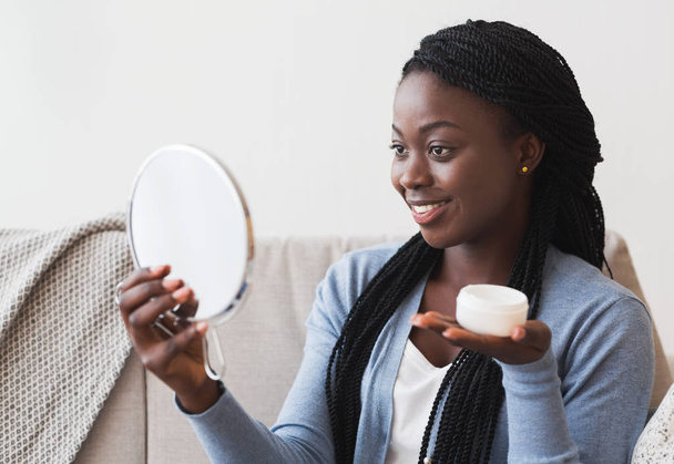Όμορφη Afro Girl Κοιτάζοντας στο μεγεθυντικό καθρέφτη και κρατώντας κρέμα Jar - Φωτογραφία, εικόνα