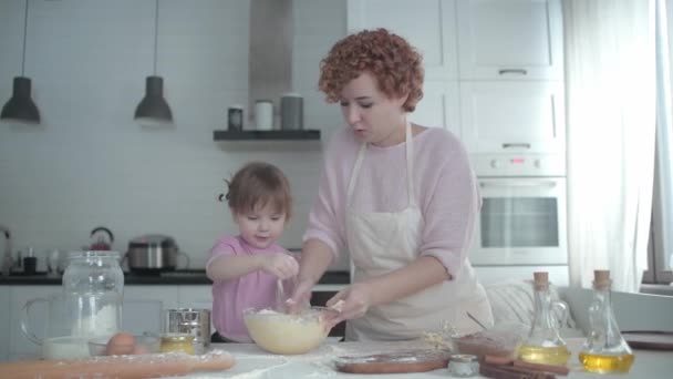 Anya a lányával a konyhában egy tányéron gyúrja a tésztát. A lány rosszalkodik a konyhában. Anya főz a kislányával. Az anya megtanítja a gyerekét főzni. Vidám főzés sütés. - Felvétel, videó