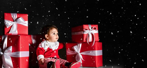 kleines Baby-Baby mit Weihnachtsmann-Kostüm mit roten Schachteln auf dunklem Hintergrund - Foto, Bild