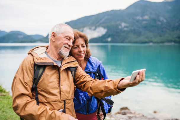 A senior pensioner couple standing by lake in nature, taking selfie. - Фото, изображение