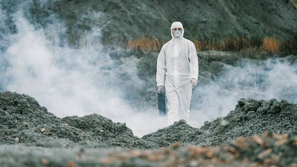 Лаборант в маске и защитном костюме, ходит по суше с ящиком для инструментов через токсичный дым - Фото, изображение