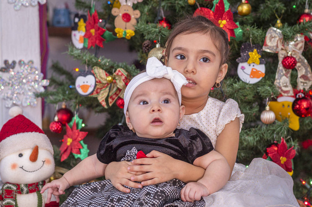 Tyttö katsoo pikkusiskoaan halaten, istuu koristellun joulukuusen vieressä, he katsovat jotain hyvin
 - Valokuva, kuva