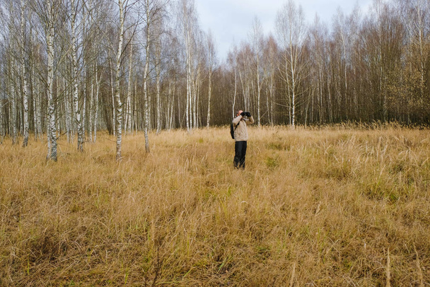 Un uomo con uno zaino guarda attraverso il binocolo. Erba gialla secca su un prato di betulla foresta. Natura ucraina. Concetto di autoisolamento. - Foto, immagini