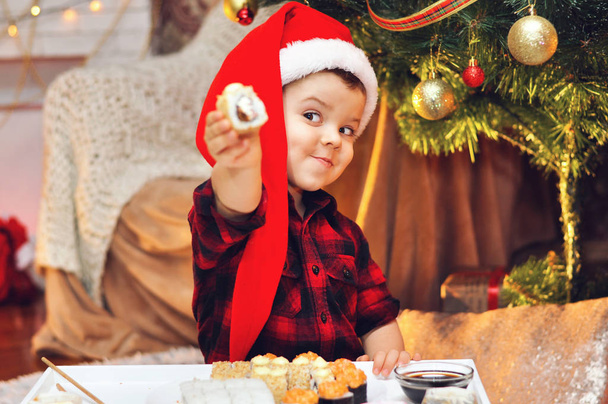 Cute boy eats sushi sitting under a Christmas tree in a red Santa hat and a red checkered shirt. - Photo, Image