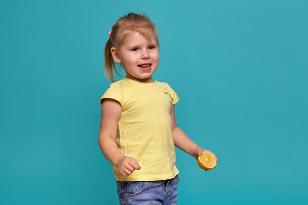 Крупный план студии с красивой маленькой девочкой. Маленькая блондинка в жёлтой футболке на синем фоне. Эмоции ребенка, который ест лимон
. - Фото, изображение