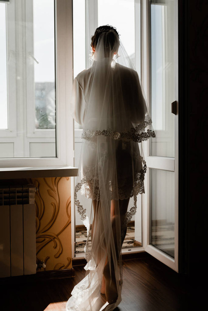 Portrait of young gorgeous bride on the balcony. Beautiful girl dresses and gathers for a wedding ceremony in apartments while waiting for the groom. Bride looking at her dress. Bride's morning - Photo, Image