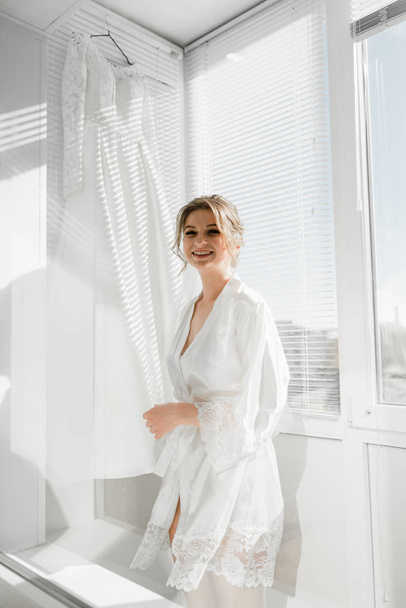 Portrait of young gorgeous bride on the balcony. Beautiful girl dresses and gathers for a wedding ceremony in apartments while waiting for the groom. Bride looking at her dress. Bride's morning - Fotografie, Obrázek