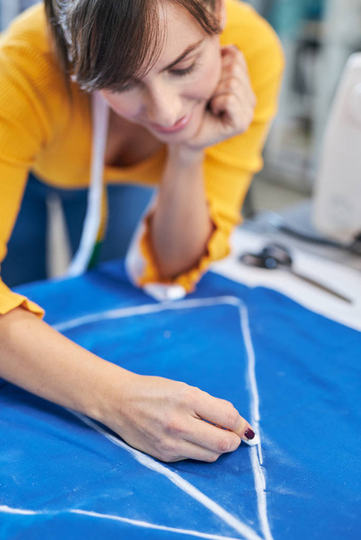 Creative caucasian fashion designer standing in her studio and drawing scheme on blue linen for a beautiful night dress. - Φωτογραφία, εικόνα