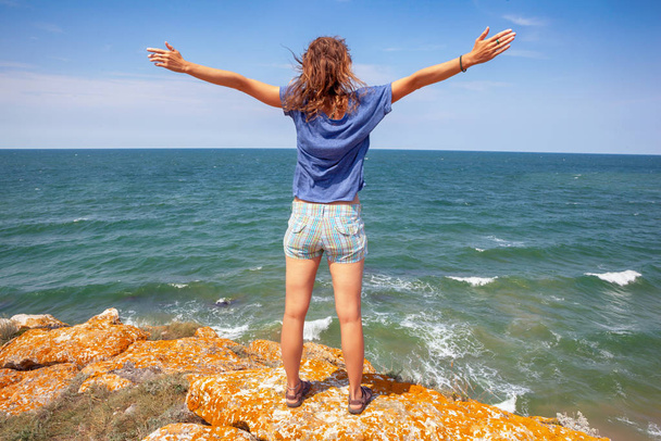 Young woman celebrating the beauty of nature standing on the rocks overlooking the sea with her arms outspread as she embraces the sun and life - 写真・画像