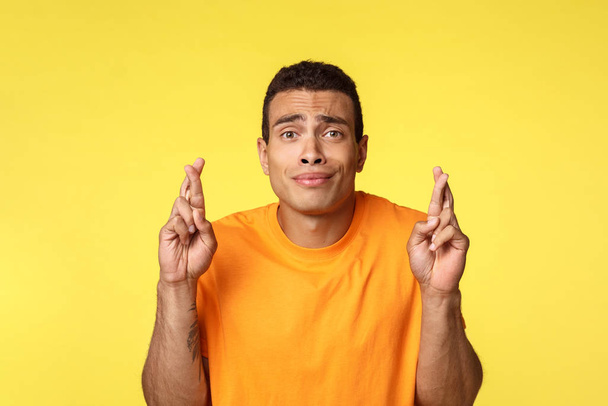 Hopeful worried young handsome man praying for wish come true, smirk silly and nervous, frowning as cant wait hear important results, want apply job, cross fingers good luck, yellow background - Fotoğraf, Görsel