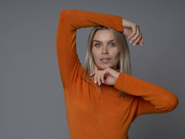 Portrait of amazing blonde woman model touching her hair and face in an orange sweater on a gray background - Foto, Bild
