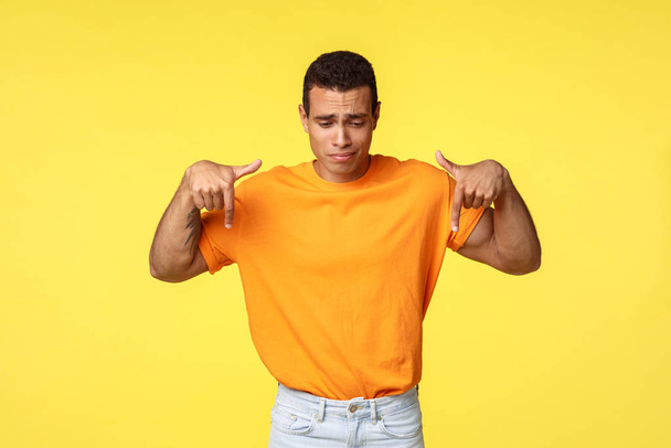 Расстроенный, разочарованный или ревнивый привлекательный молодой мужчина, спортивное тело, оранжевая футболка и белые брюки, хмурясь от сожаления или грусти, указывая вниз, хмурясь неспокойно, желтый фон
 - Фото, изображение
