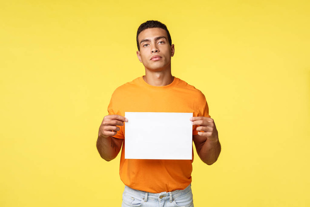 seriös aussehender motivierter und selbstbewusster junger Mann in orangefarbenem T-Shirt, leeres Papier in der Hand und kamerasicher, Werbung für etwas Geschriebenes, Werbung für Konzept, gelber Hintergrund - Foto, Bild
