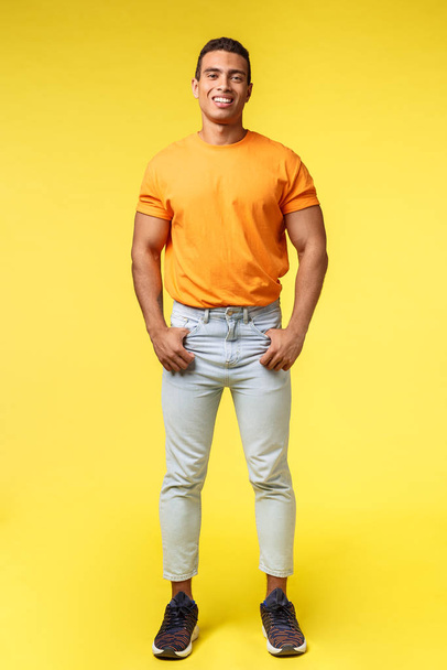 Vertical full-length studio shot young hipster guy with mascular body, standing orange t-shirt and white pants, hold hands in pockets, smiling camera pleased and confident, casual pose - Photo, Image