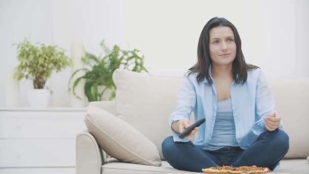 Slowmo. A young woman is switching on the TV programm, while sitting on a sofa. She is eating pizza with surpised facial expression. Yummy-yummy. 4K. - Video
