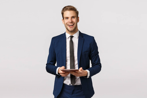 fröhlicher, eleganter bärtiger Geschäftsmann in klassischem Anzug und Krawatte, mit digitalem Tablet lachend und enthusiastisch lächelnd, mit Businessplan geschriebenem Gerät, vor weißem Hintergrund stehender Rede - Foto, Bild