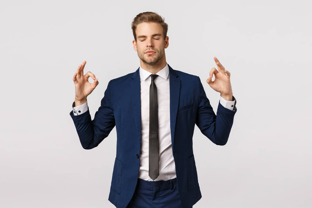 Dont panic, taking emotions under control. Peaceful and patient good-looking blond businessman in classic suit, make zen sign, spread hands sideways, close eyes, meditating, reliese stress - Photo, Image