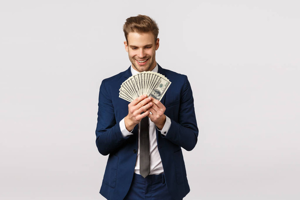 Feliz y emocionado, hombre de negocios guapo en traje clásico, contando dinero, sosteniendo grandes dólares en efectivo y sonriendo con expresión complacida, encantada, de pie fondo blanco contento
 - Foto, imagen