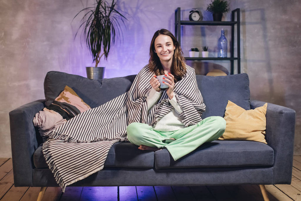 Een jonge vrouw zit op een bank in een gezellige kamer gewikkeld in een deken, thee drinken en tv kijken. - Foto, afbeelding
