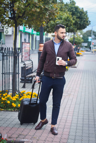 Handsome man with a suitcase for tourism. Exit the house for travel. Fashionably dressed guy and coffee. Stock Photo. - Foto, afbeelding